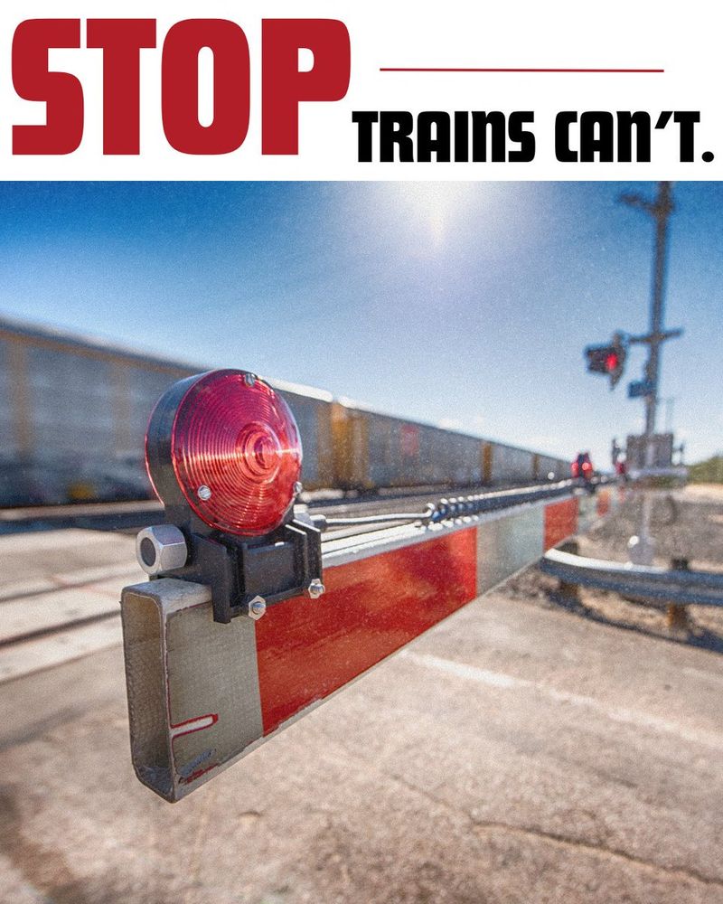 STOP. Trains can't." Close-up view of a red, illuminated railroad crossing signal arm, with a blurred train and additional crossing signal in the background under a clear sky.