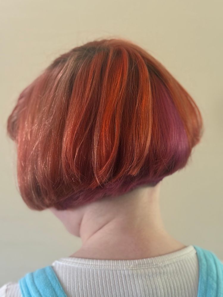 A young person looks to the wall to show off the back of their new hairdo. I cut and colored it at home, I am not a professional but I’m proud! The bob features layers of hot pink, orange, and purple. 