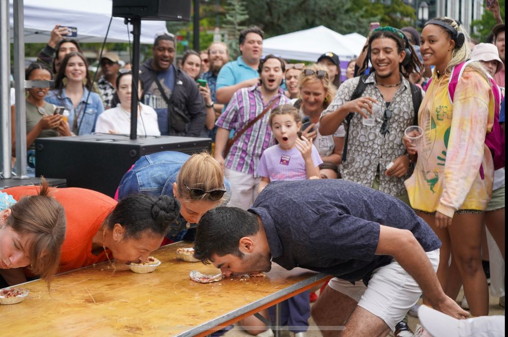 Four people bent over a table eating mini pies with their hands behind their backs, while a crowd looks on in awe, delight, and horror