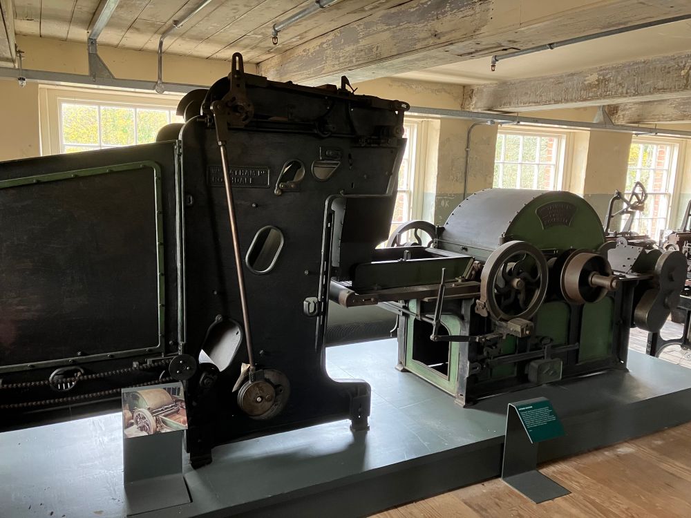 A large green and black machine with moving rods and wheels sots on a green base in a room with windows behind and cream coloured walls and ceiling. 