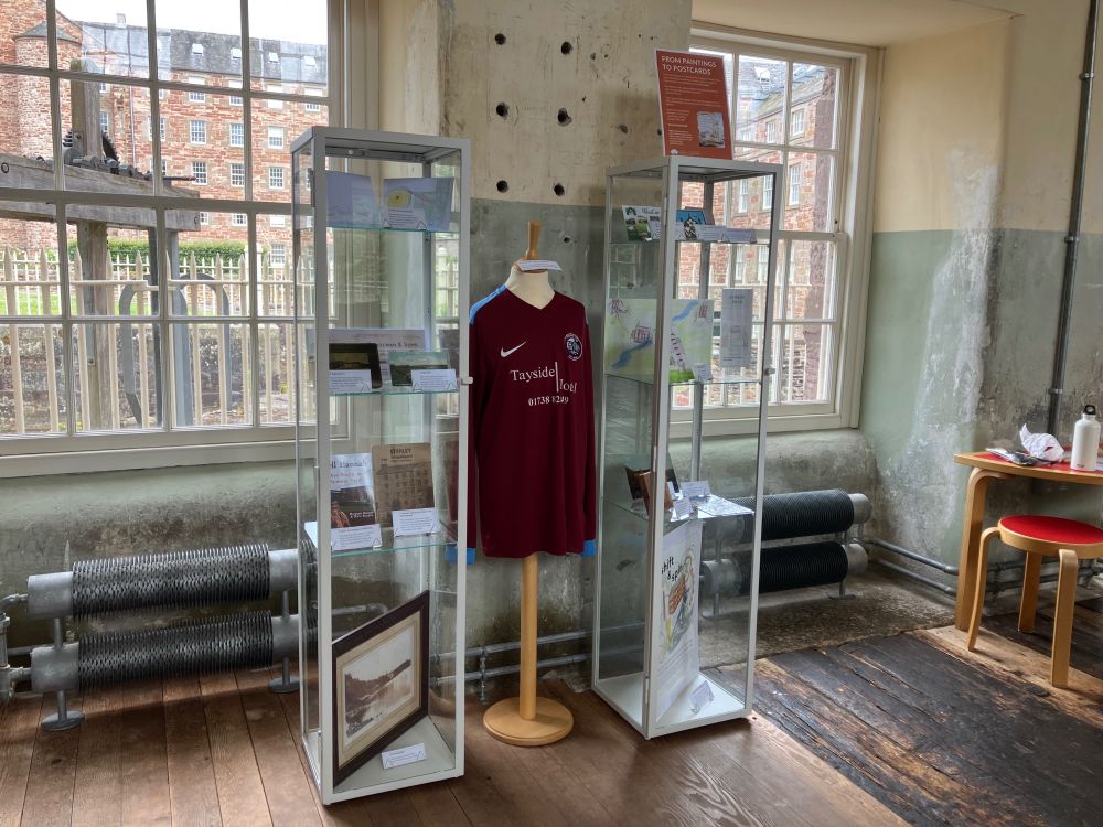 Two tall glass cabinets containing a range of objects with windows behind them. A torso mannequin with a maroon football shirt on it stands between them.