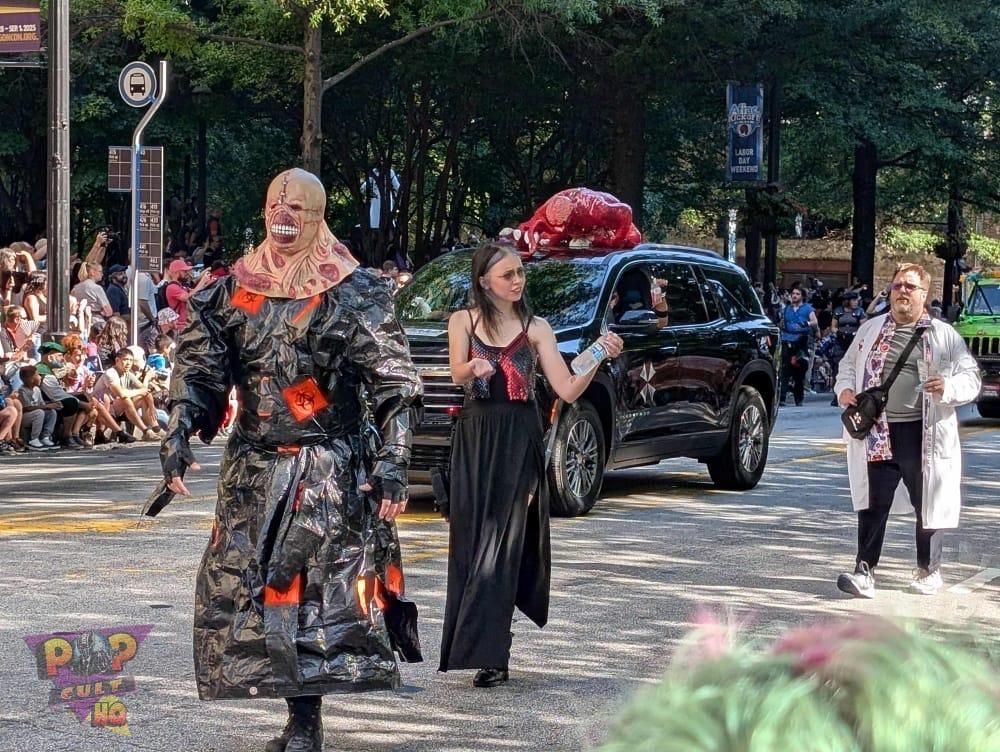 Nemesis, a young lady in a black and red dress, and a man in a white lab coat parade while flanking a black SUV with a red monster on the roof.