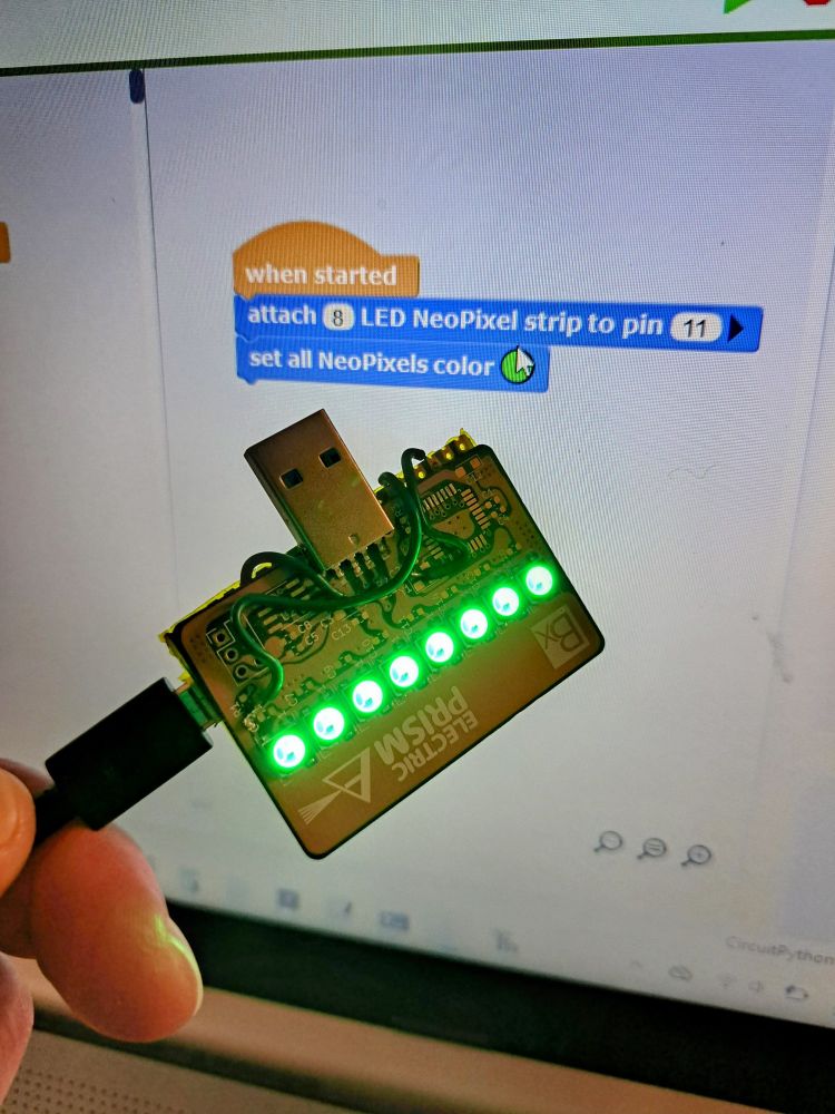 A picture of a bodged together PCB with neo pixels being driven by a raspberry pi pico. The device is held in front of a laptop screen which displays the MicroBlocks code that is being run on the board.