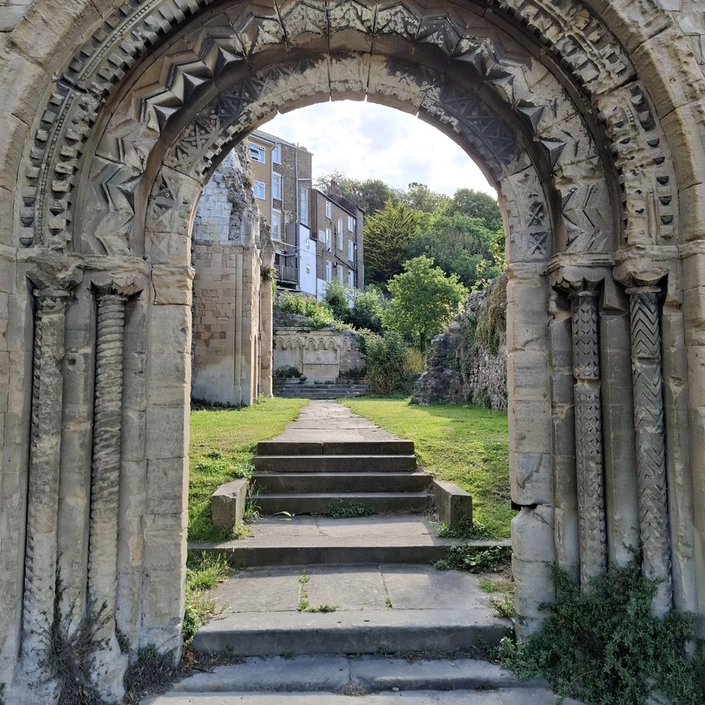 A colour photograph of the West entrance to St James' church Dover. The photo shows the arch to the doorway with Norman chevron "zig-zag" patterns. The picture shows the aisle leading to the far end with remains of the internal walls. Either side of the aisle is surrounded by grass. 