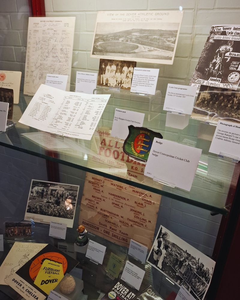  An angled shot of an exhibition display case showing ephemera from sports teams and events at Dover Athletic Ground (Crabble Athletic Ground), including programmes, poster, a cricket club badge, sepia and black and white photographs of sports teams