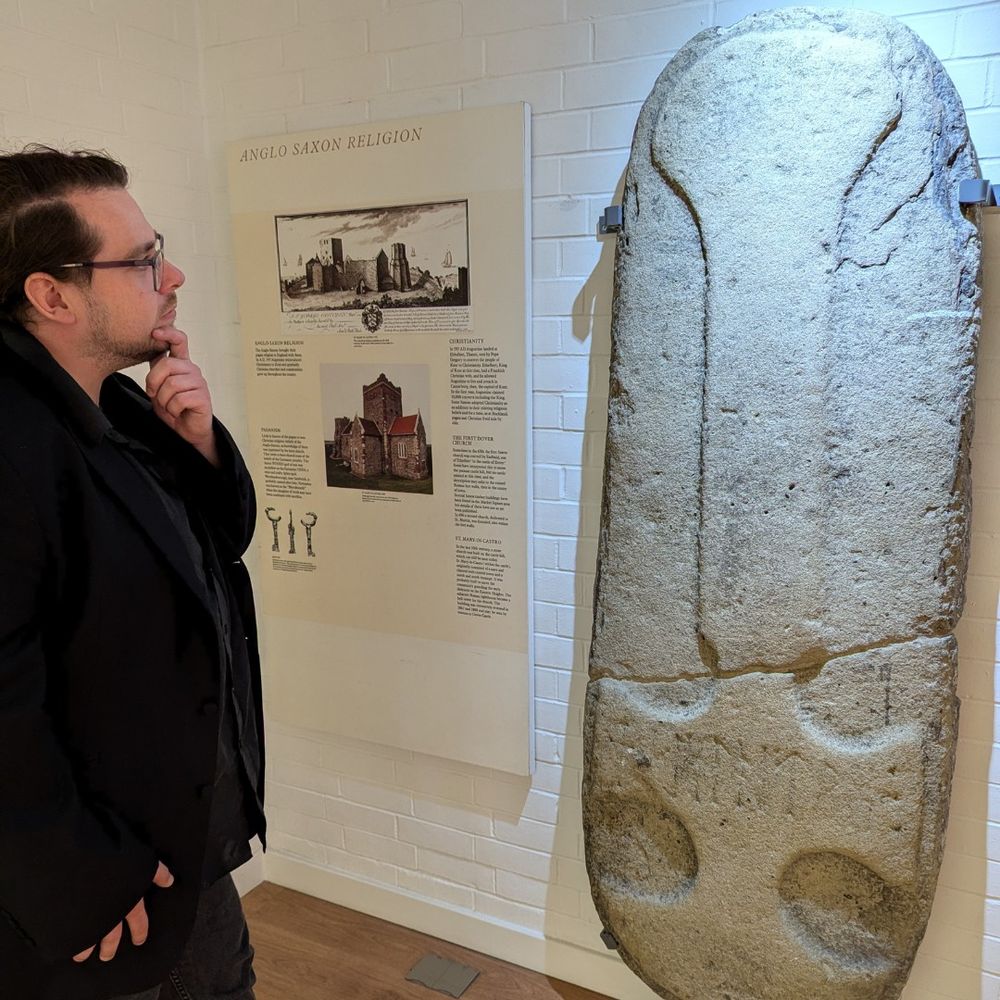 Curating Visibility fellow Karl. Karl has dark brown hair and wears glasses. He is wearing a black jacket. He is looking at a large Anglo-Saxon grave slab featuring a cross with runes that has been mounted on a wall. 