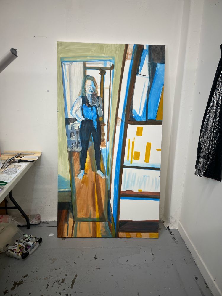 Studio corner with white walls and gray floor. There is a large canvas leaning against the wall straight ahead depicting an under painting in browns, blues and blacks. On the right is a sequined garment on the wall on the left is a table and under the table a bin of oil paint tubes