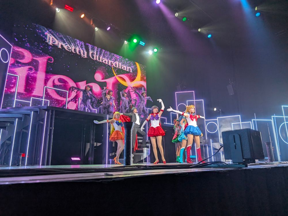 Wide view of the stage from Sailor Moon: The Super Live that shows the entire cast - all five of the Sailor Scouts, Tuxedo Mask, Queen Beryl, and the backup dancers/henchwomen.