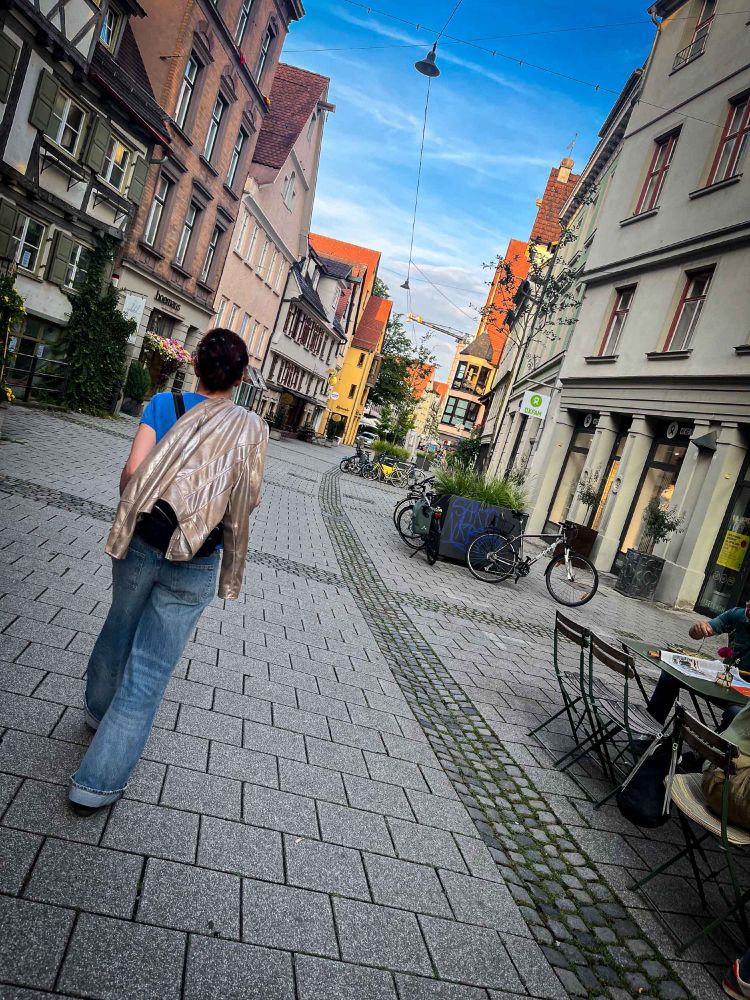 Fee von hinten, sie schlendert durch eine Altstadtgasse, eine Jacke über der Schulter, über allem blauer Himmel.