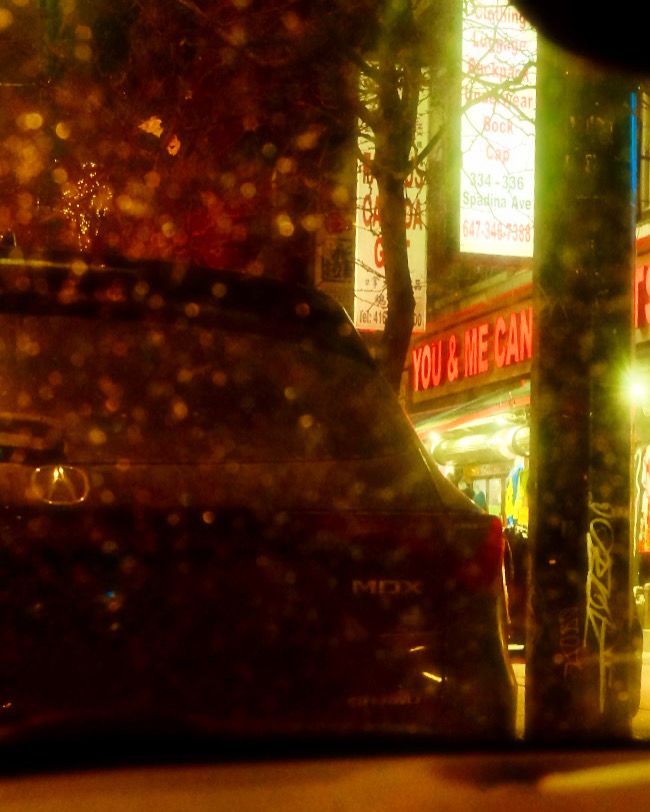 Evening view from inside a parked car, looking out at a partially hidden storefront sign that reads ‘You and Me Can.’ A dark parked car and a lamppost frame the scene, while streetlights cast a moody glow. Light snow falls on the windshield, softly diffusing the lights.