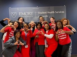 A group of 12 Emory Student Health staff members wearing red for Heart Health and making a heart shape with their hands. They are standing in front of a banner that reads: "Emory University Student Health Services."