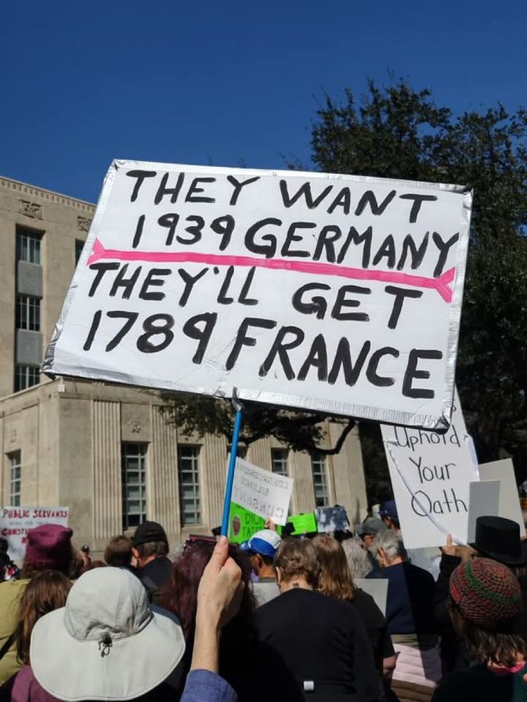Protestors holding signs, the most prominent one reads: "The want 1939 Germany. They'll get 1789 France."