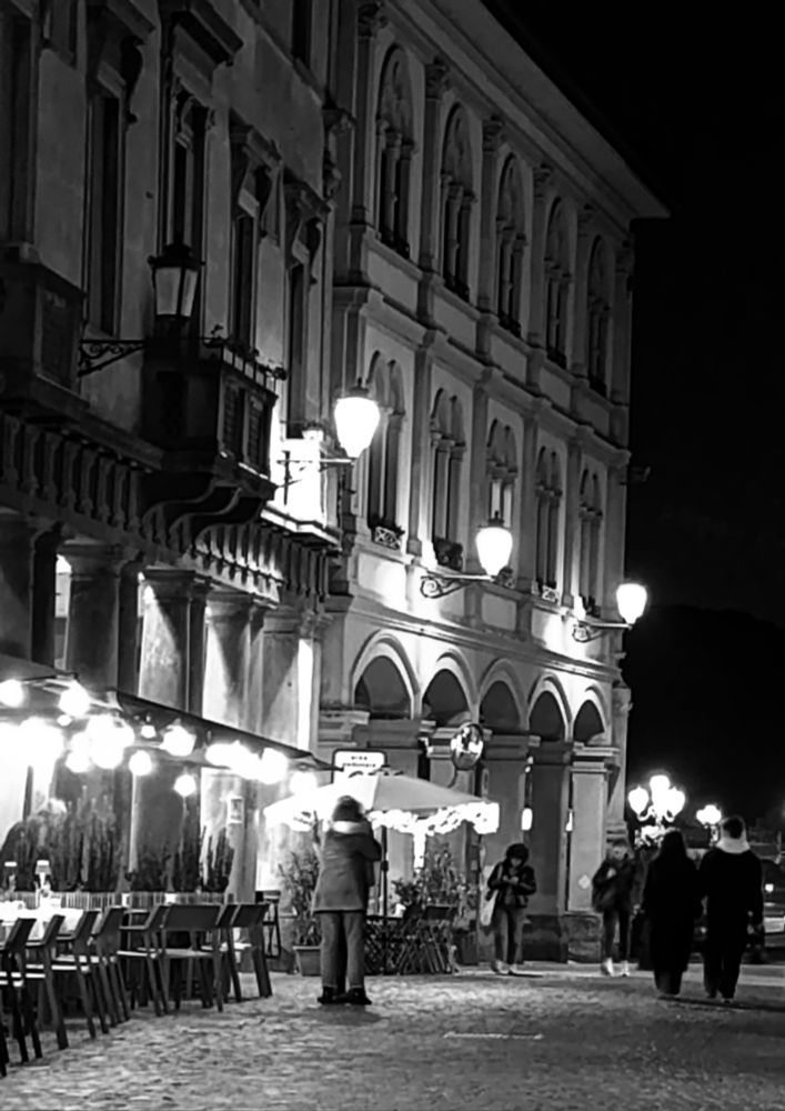 Due ragazzi si abbracciano per strada, di sera. Sul fianco destro alcuni passanti. Alcune luci illuminano la strada, costeggiata da portici e tavolini, sulla sinistra