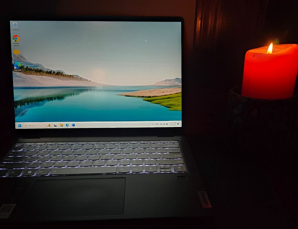 Open laptop with nature background scene and slightly backlit keyboard sitting in the darkness beside a lit candle.  Can't see much else.