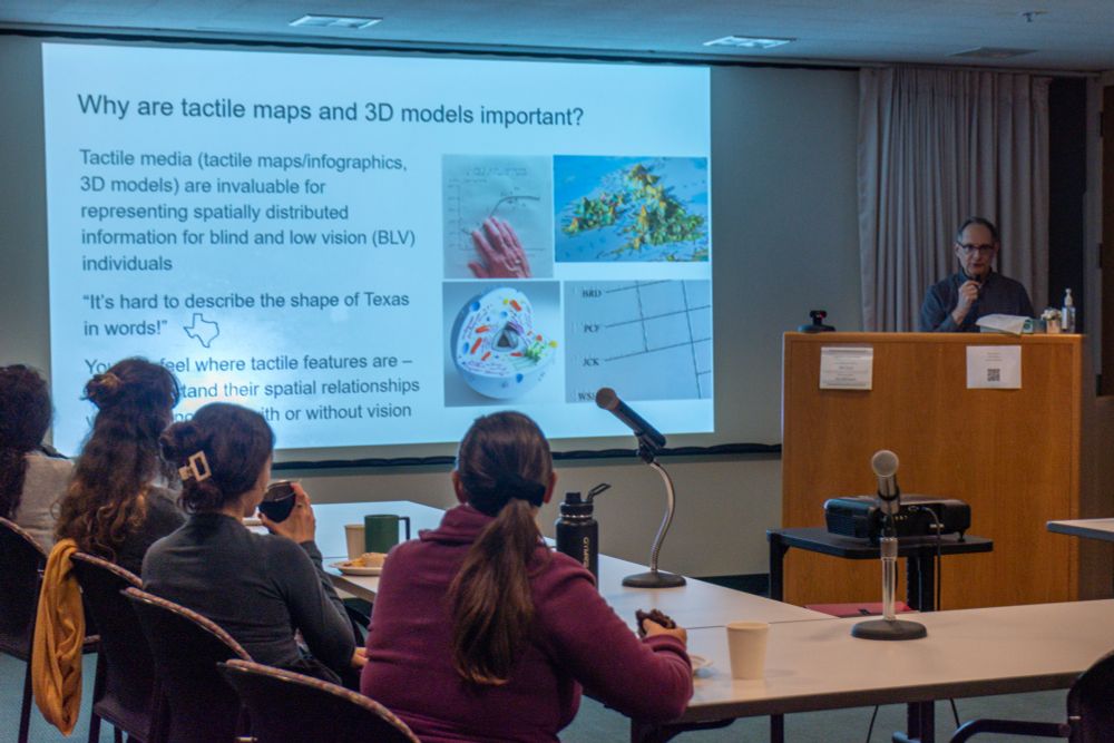 A speaker at a podium presents a slide titled “Why are tactile maps and 3D models important?” to an audience seated at tables. The slide includes images of tactile graphics and raised maps.