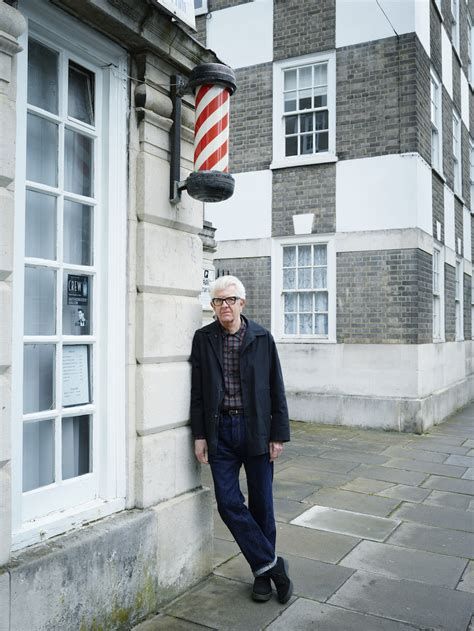 Nick Lowe in turned-up jeans, standing on a street corner; above him is a red and white striped barber's pole. Photo Dan Burn-Forti.