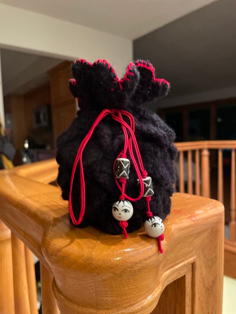 A black yarn dice bag crocheted using a crocodile stitch has a red embroidered edge with a red string tie with black and white ceramic beads