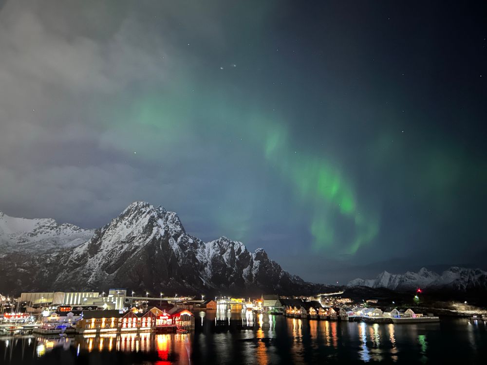In the foreground is the colourful lights of a town reflected in the Fjord water. There is a snowy mountain behind the town, and in the sky can be seen the green Aurora.