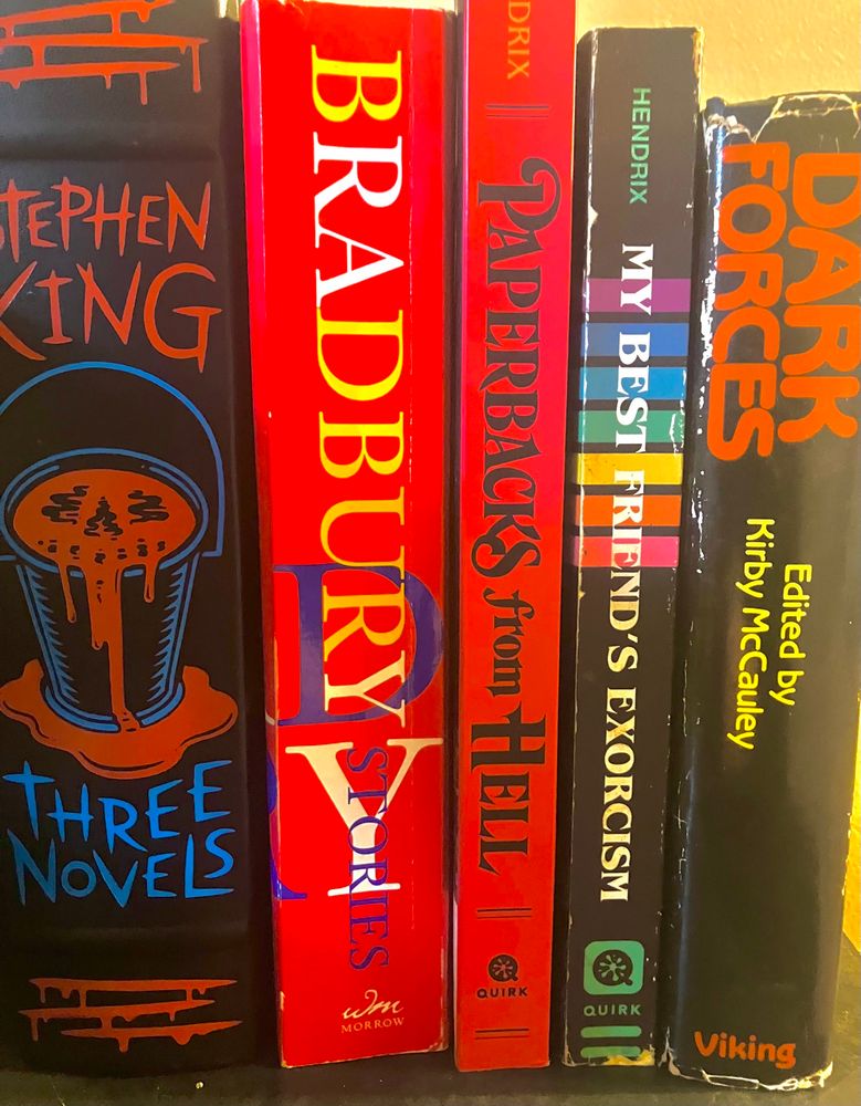 Books on a shelf: Carrie/Salme’s Lot/The Shining by Stephen King, Stories by Ray Bradbury, Paperbacks from Hell by Grady Hendrix, My Best Friend’s Exorcism by Grady Hendrix, and Dark Forces edited by Kirby McCauley