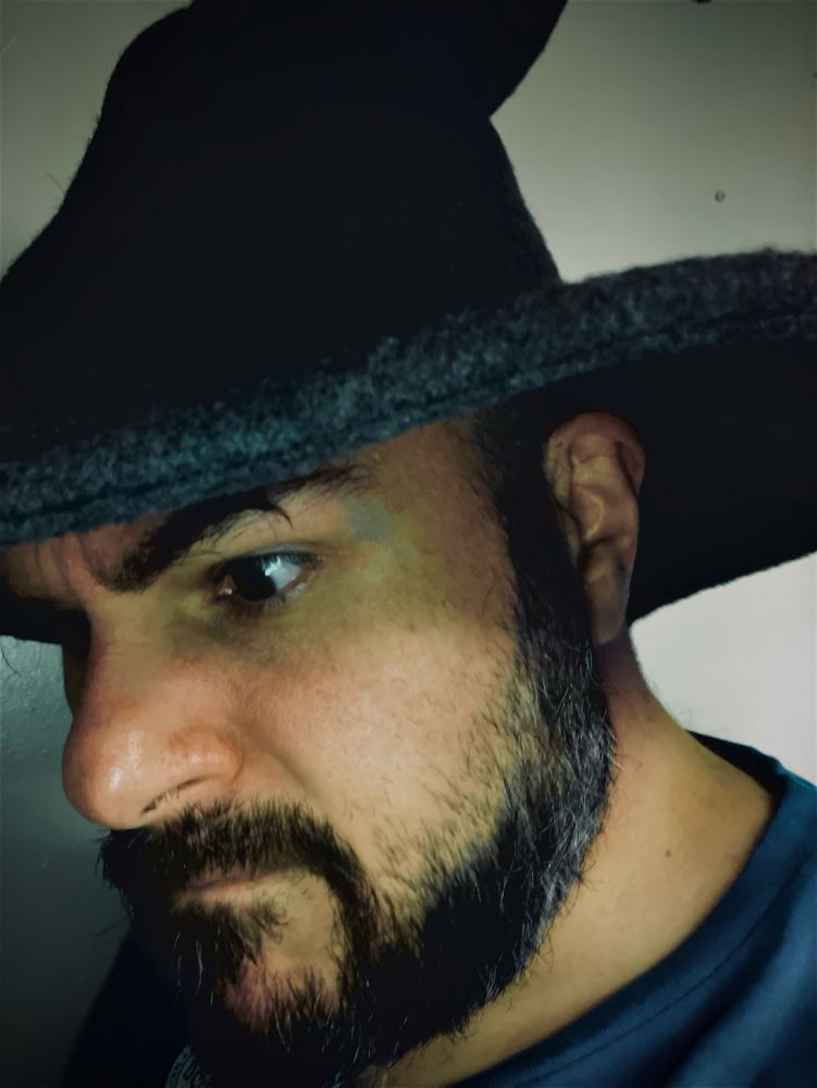 A photo of me in a wizard hat. It’s a side profile. My nose is very pronounced, I have bushy eyebrows and a dark beard. The hat is gray but looks almost too dark to see. 