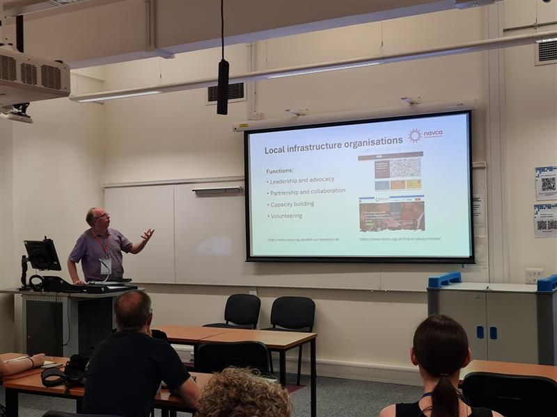 A presenter stands in front of a computer desk in front of an audience seated in rows. On the large screen next to the presenter is a slide from their presentation