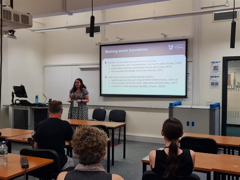 A presenter stands at the front of a room, in front of an audience seated in rows. On the large screen next to the presenter is a slide from their presentation