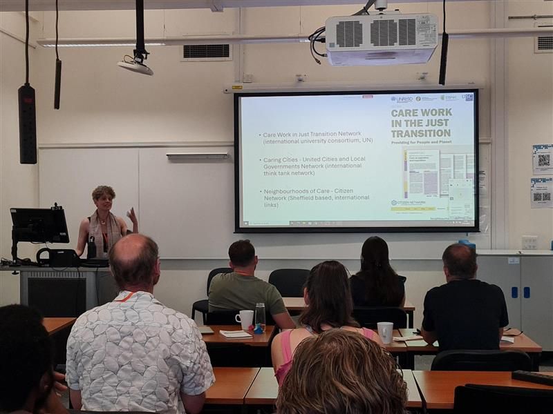 A presenter stands in front of a computer desk in front of an audience seated in rows. On the large screen next to the presenter is a slide from the presentation