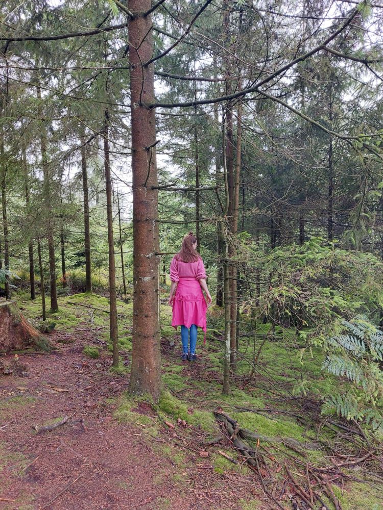 Photo of a woman standing in the forest, wearing a pink sweater and a pink raincoat and blue jeans