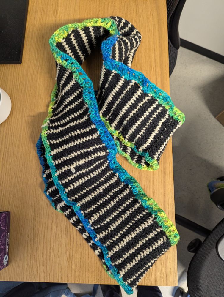 An infinity scarf on a desk. The scarf has a black and off-white strip pattern and a border that shifts between blue, green, and yellow 