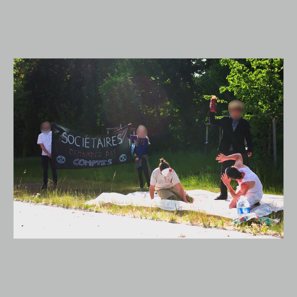 Deux personnes allongées au sol, tachées de faux sang, une personne tenant une bouteille pleine de faux sang, et deux personnes tenant une banderole "Sociétaires, demandez des comptes !".