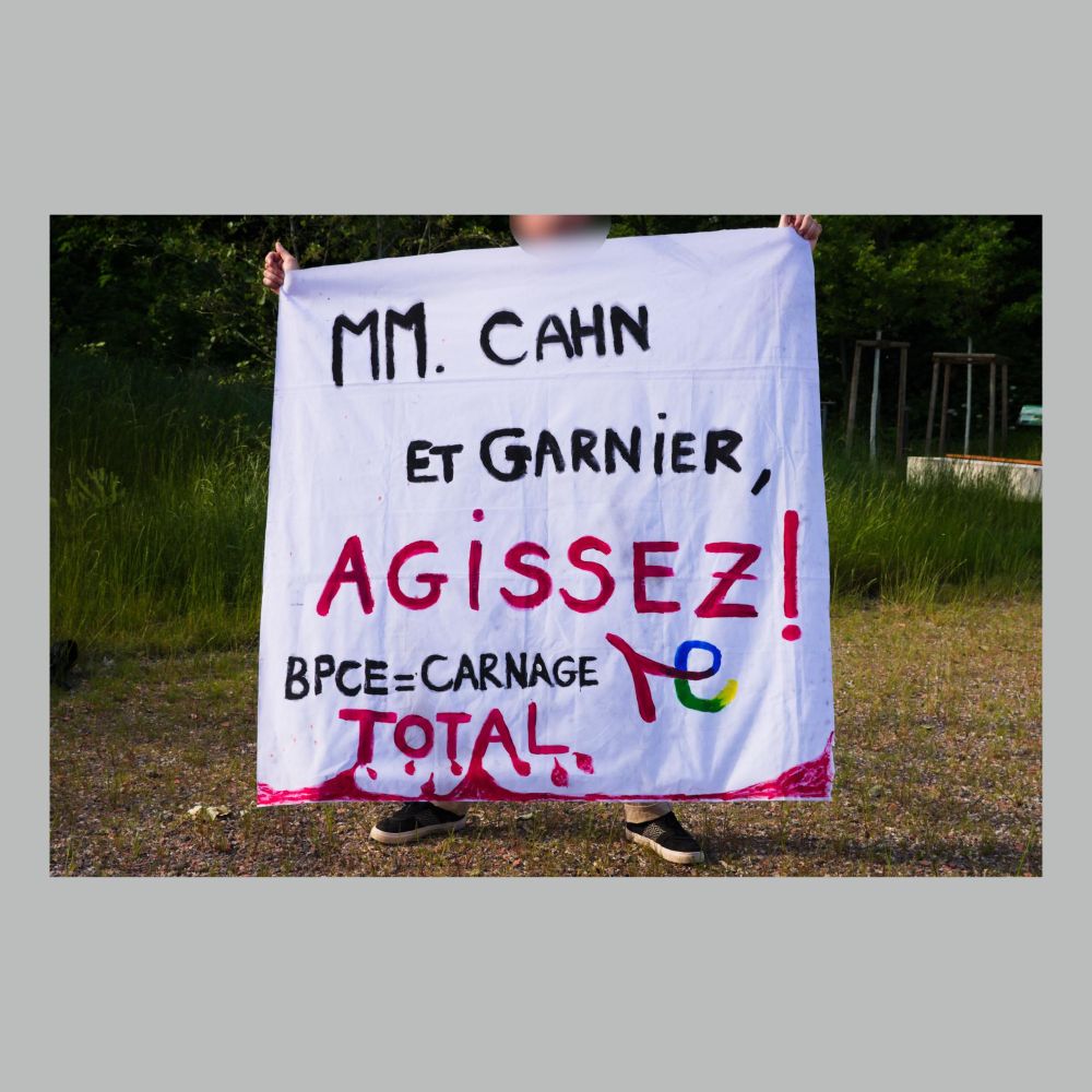 Personne tenant une banderole "MM. Cahn et Garnier, AGISSEZ ! BPCE = carnage Total"
