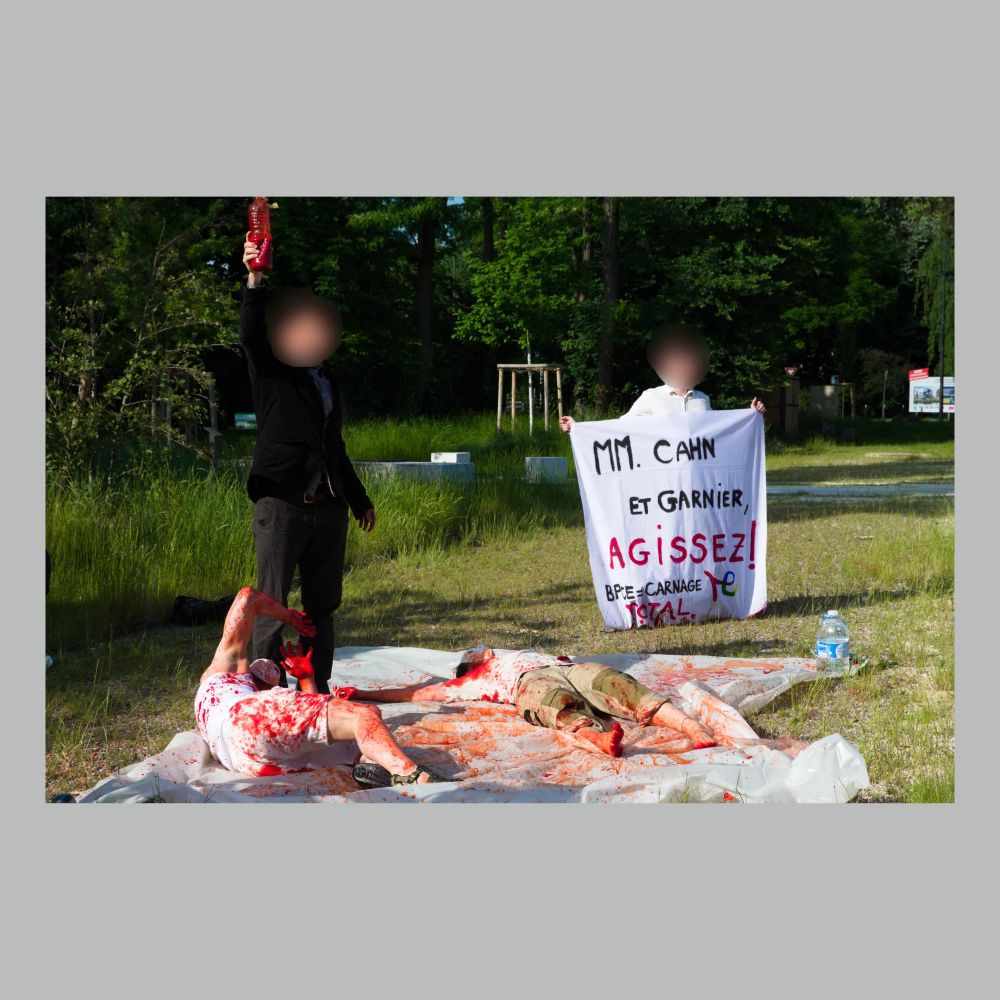 Deux personnes allongées au sol, tachées de faux sang, une autre brandissant une bouteille pleine de faux sang, et une dernière dans le fond qui tient une banderole "MM. Cahn et Garnier, AGISSEZ ! BPCE = carnage Total"
