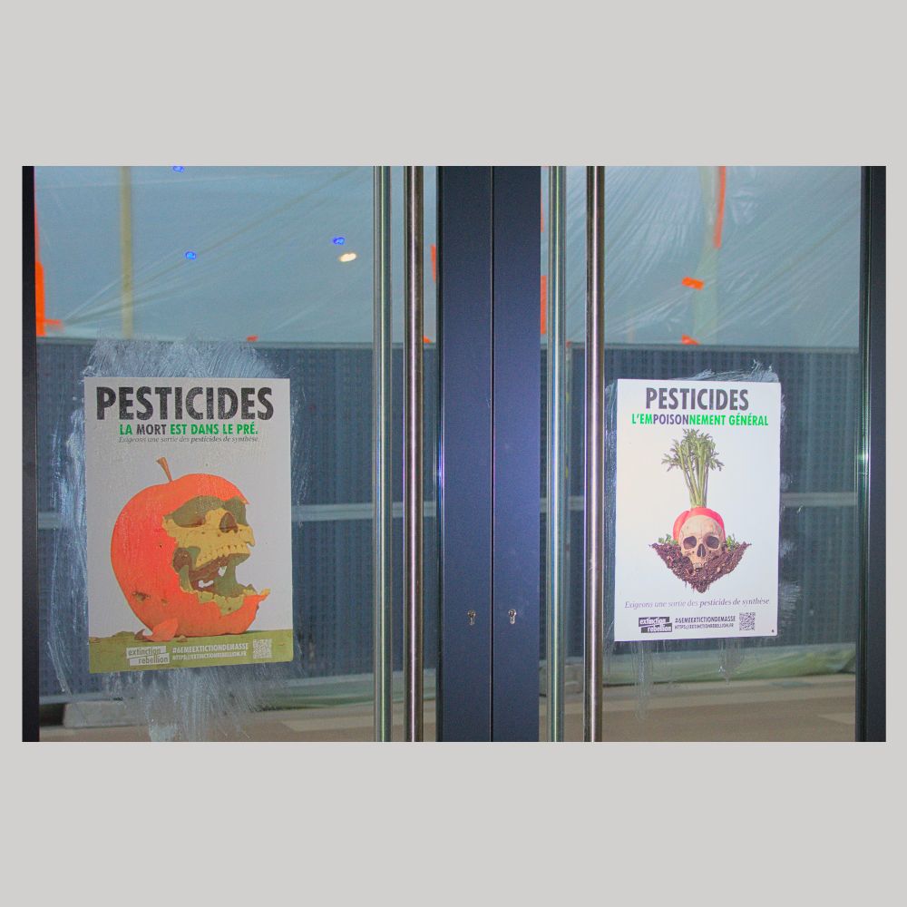 Photo de deux affiches "Pesticides, la mort est dans le pré" et "Pesticides, l'empoisonnement général" sur une porte vitrée à double battant. 