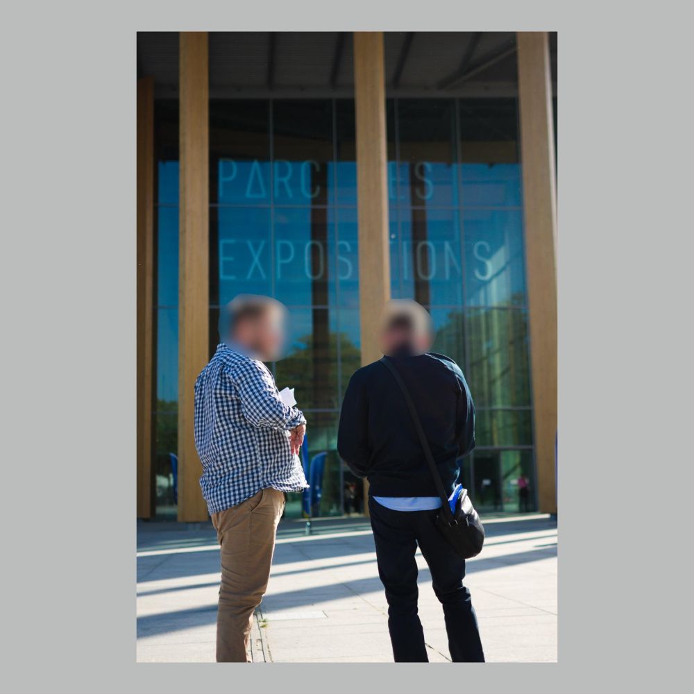 Deux personnes discutant, l'une ayant des tracts. Dans le fond, un grand bâtiment en verre : Parc des Expositions.
