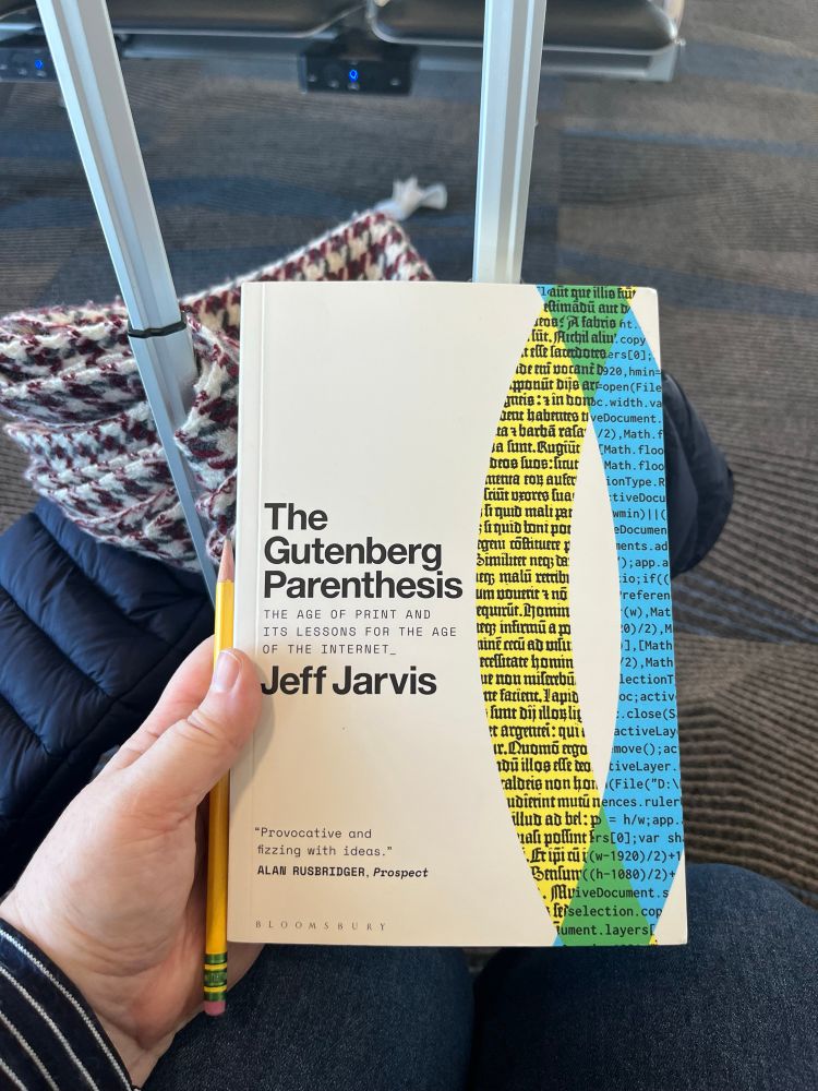 A roller bag, puffer vest, and houndstooth scarf provide the backdrop to a copy of Jeff Jarvis’s The Gutenberg Parenthesis, held by a white-skinned hand. Also present is a pencil for notes (of course!)