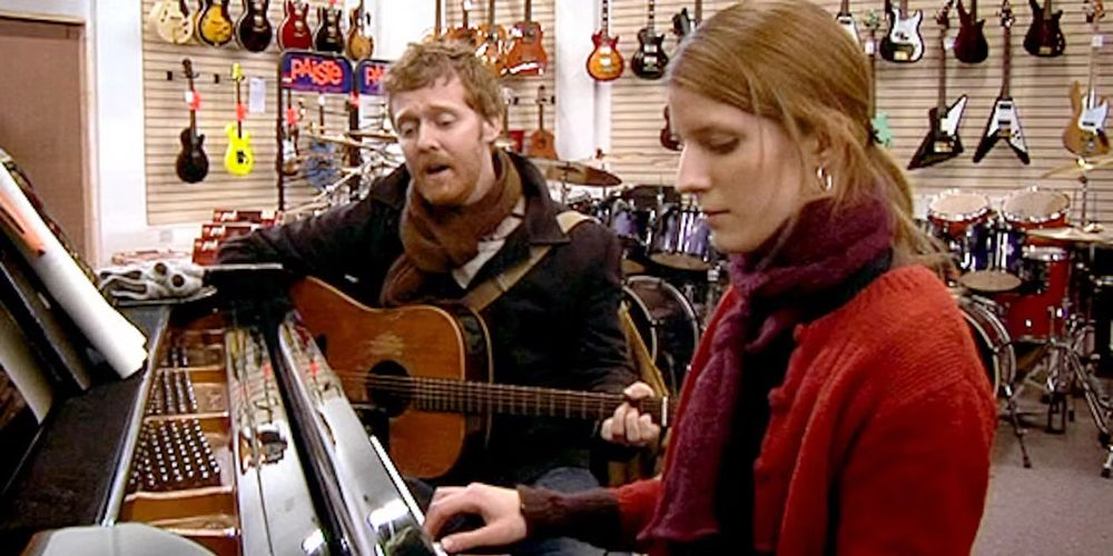 It’s Glen Hansard and Marketa Irglova sitting in a music store. He’s holding a guitar and she’s at a piano. He’s got a scarf on and she’s got a scarf on. He’s got a dark coat and she’s wearing a red sweater. There’s lots of electric guitars on hangers on the wall, and drums in the back.