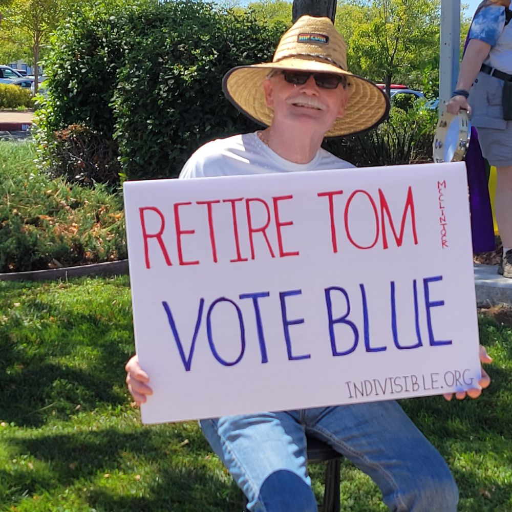 Man smiling, holding sign that says 'Retire Tom McClintock - VOTE BLUE - indivisible.org'