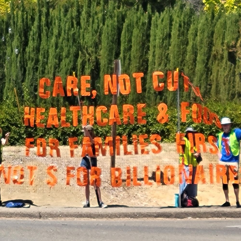 Protesters holding up large sign that says "Care, not cuts to healthcare & food for families first not $ for billionaires" - standing on corner in front of trees.