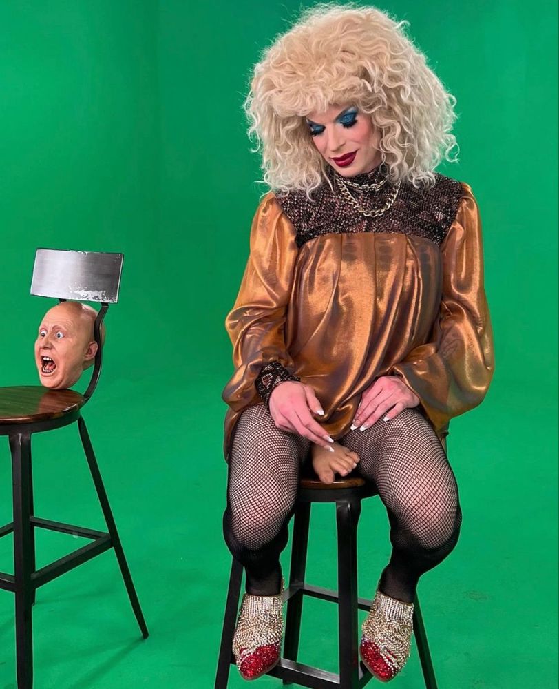 Katya Zamolodchikova sitting on a stool on the set of UNHhhh with a fake foot between her legs. 