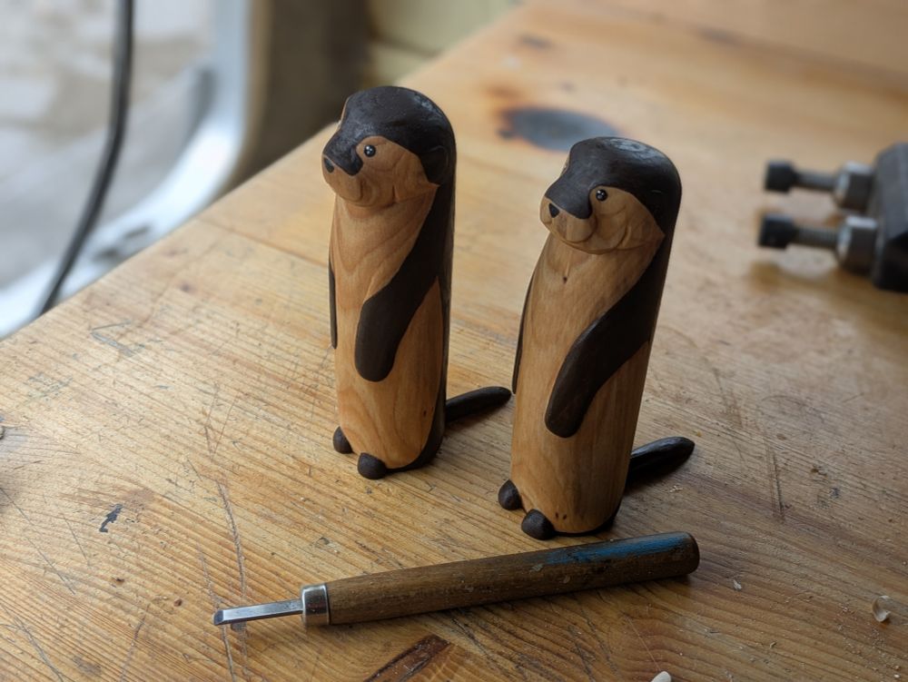 Two woodcarved otter on a workbench, with a carving tool at their feet