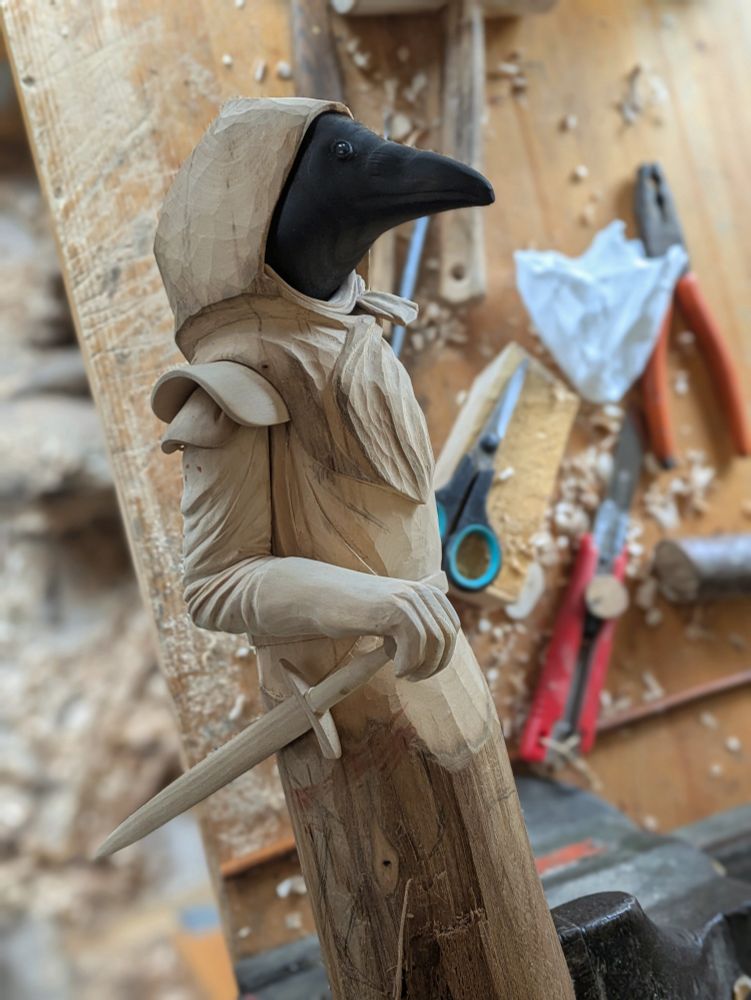 Draft of a woodcarved humanoid crow wearing a hooded cloak, shoulder armor. Its left hand rest on the handle of its sword. The arm has a more detailed clothing texture than before.