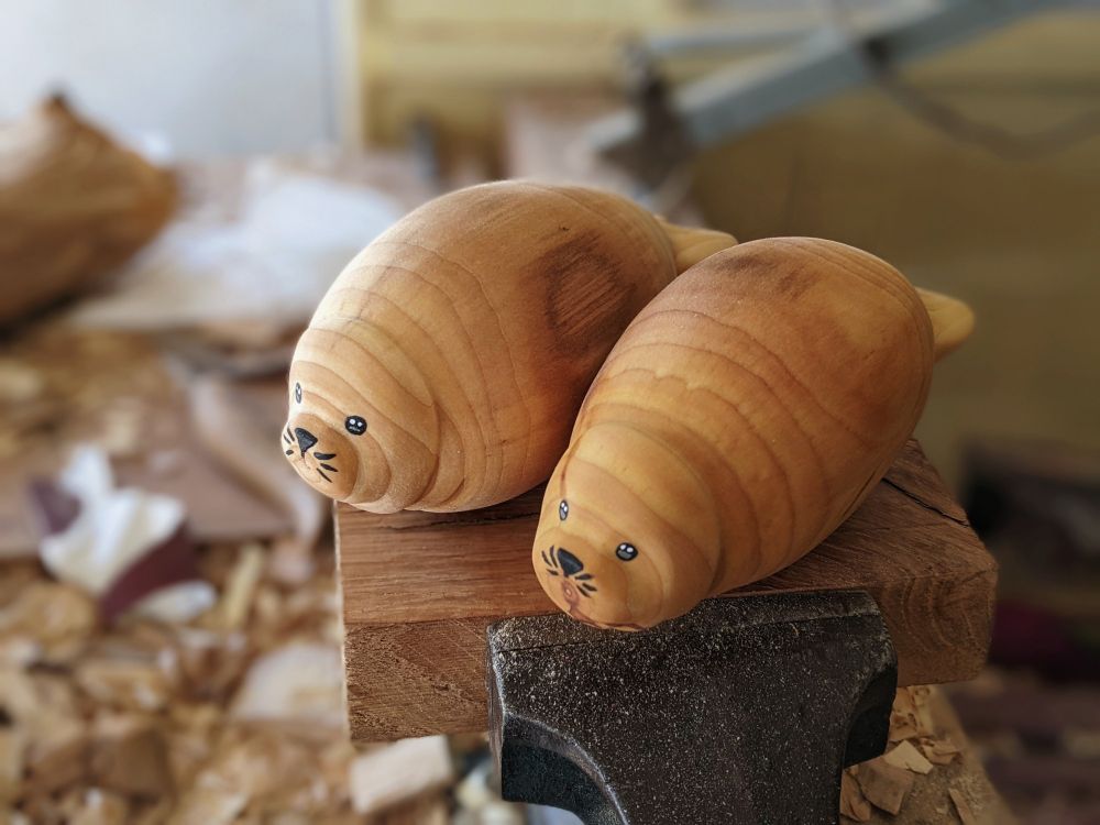 Deux phoques dodus en bois. Ils sont en postier allongés. Ils ont des bourrelets et sont dorés comme des viennoiseries. Ils sont posé sur une planche prise fans un étau. Dans le flou inyplan de travail bordélique.