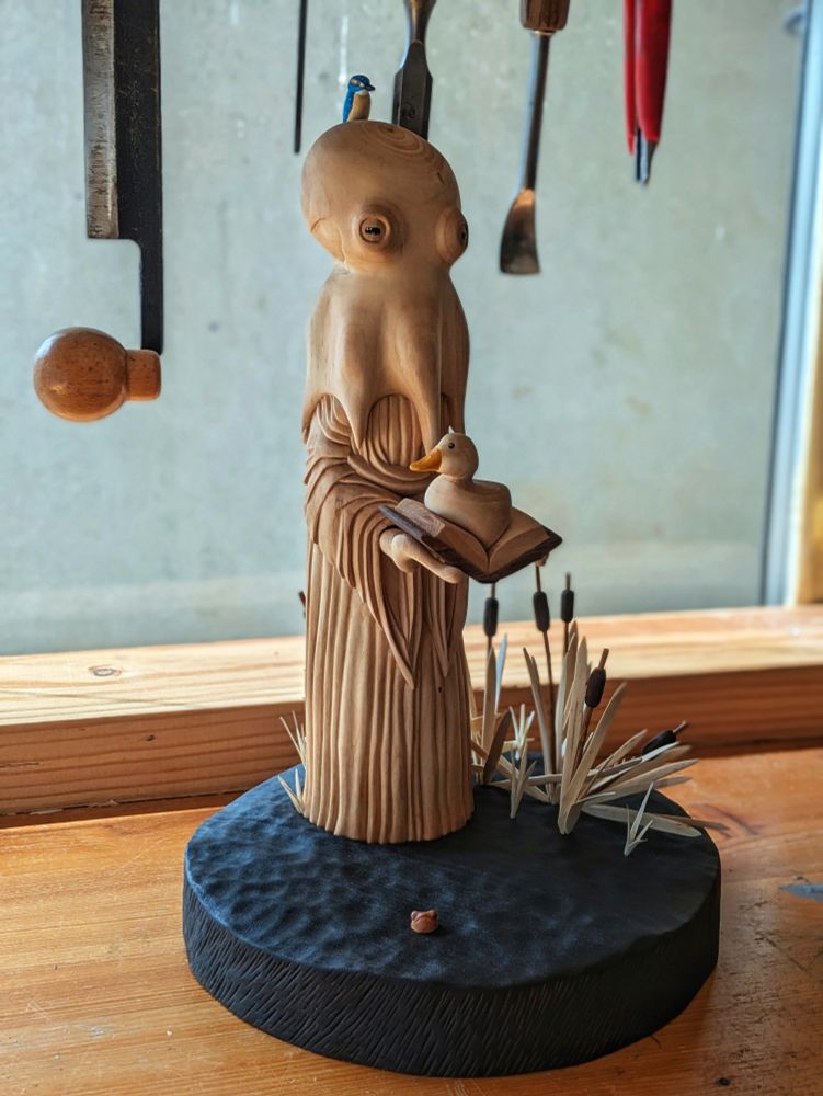 Woodcarved humanoid octopus wearing a dress, holding a book with a duck on it. It has a kingfisher on it's head. The base is a dark pond, there is a tiny head of an unhappy frog and some bulrushes