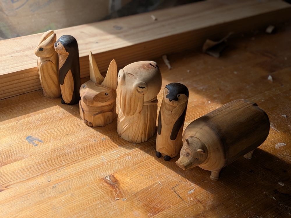 Set of woodcarvings on a workbench. From left to right : A draconid, an otter, a bat, a cuttlefish, another otter and a plump pig