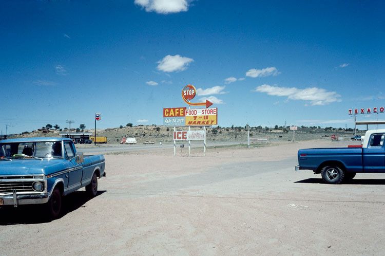Two gas stations and a roadside cafe and food store sign in the desert with two blue pickup trucks in the foreground and blue sky with a few white clouds above.