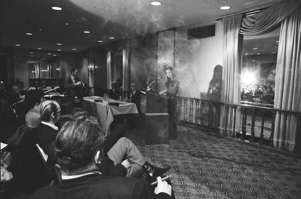 A woman stands at a podium speaking in a hotel ballroom as a small crowd of men sit in the audience listening. Smoke clouds the room, obscuring the view of the woman, as one man in the foreground grasps a cigar. 