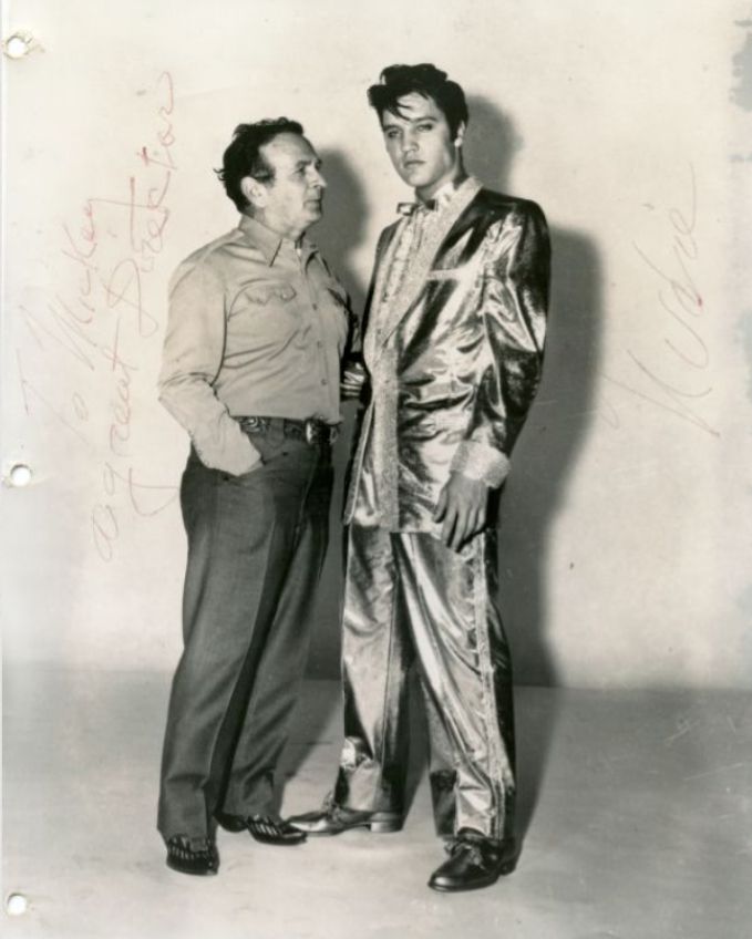 A middle aged man stands next to and facing a young man who is wearing a shiny suit and bow tie.
