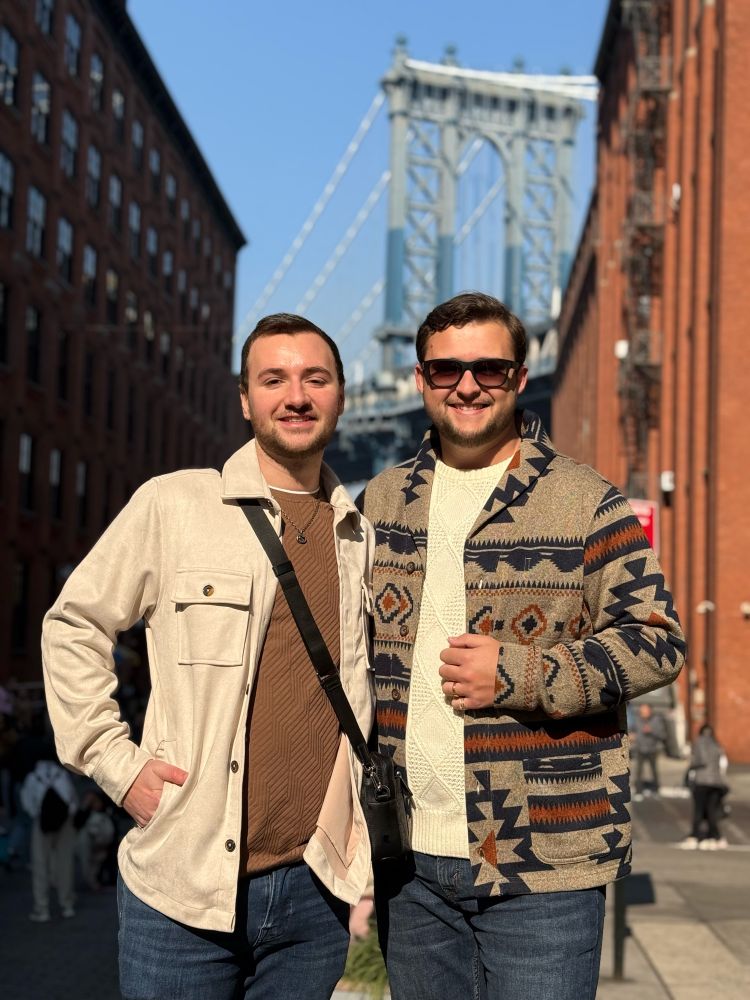 my fiancé and i smiling at the camera during morning Golden Hour, with a view of the Manhattan Bridge in the background, in between two Red Stone buildings. 