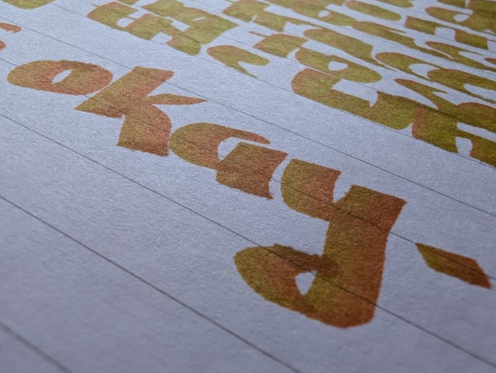 An even closer shot of my work which shows the word "okay." The ink is mainly orange with a tinge of green. The letters are chunky as the nib was held horizontally. There are a few more words which are in the top right hand corner, but they are not the focus of the photo. It is on lined paper and the translucent letters are pleasing to the eye. Diffuse lighting comes from the upper left quadrant.