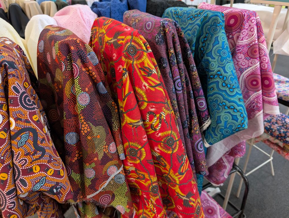 Angled shot of six rolls of fabric which are in bright and colourful original designs. From left to right roll one is okay. Yellow and black with a honeypot and and little people design. Roll two is chocolate brown with blue, red and green designs of water holes. Roll three is a bright red background with water holes and rivers and various animals printed in black and yellow. Roll four is a purple colour with pink lilac and yellow accents and symbols of gathering spots with people. Roll five is an ocean blue colour and printed in turquoise and lighter blue are footprints symbols for people and water holes. The last roll is a musk pink and is printed with white and other shades of pink featuring water holes and animal tracks. There are other fabrics and racks scattered around in the background indicating that this is a fabric store.